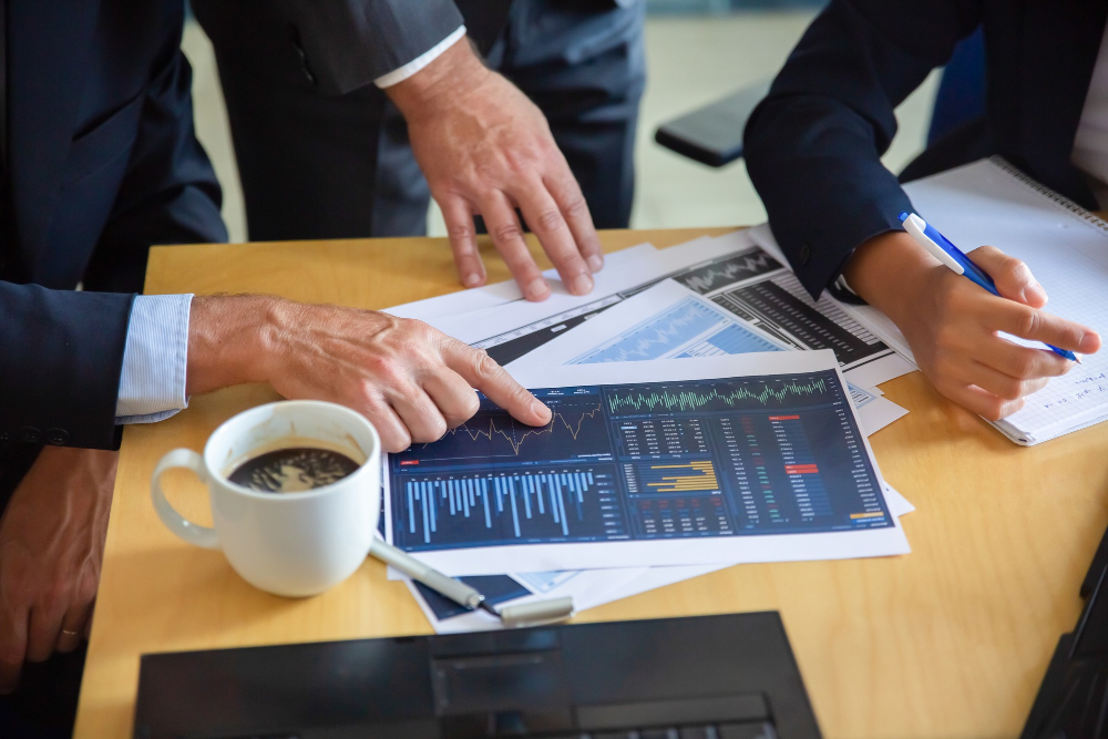Business professionals review financial charts and market data at a desk while one points to a report and another takes notes.