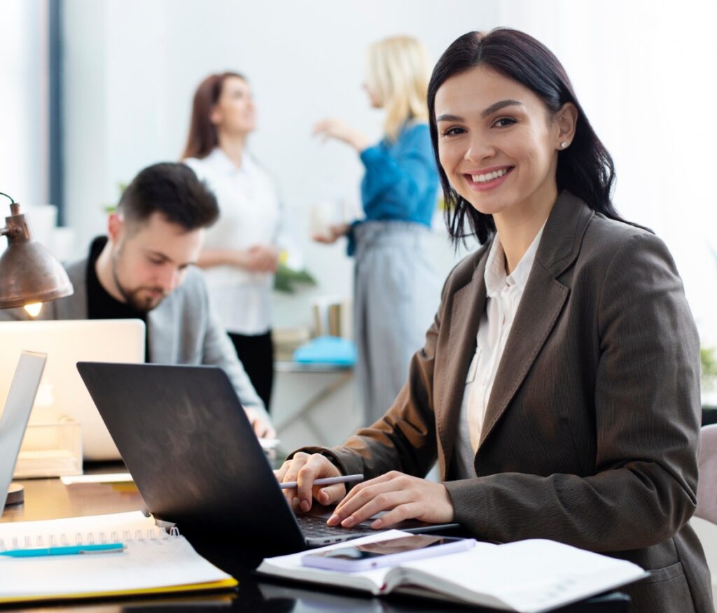 A professional smiles at the camera while working on a laptop in a modern office, with coworkers collaborating in the background.