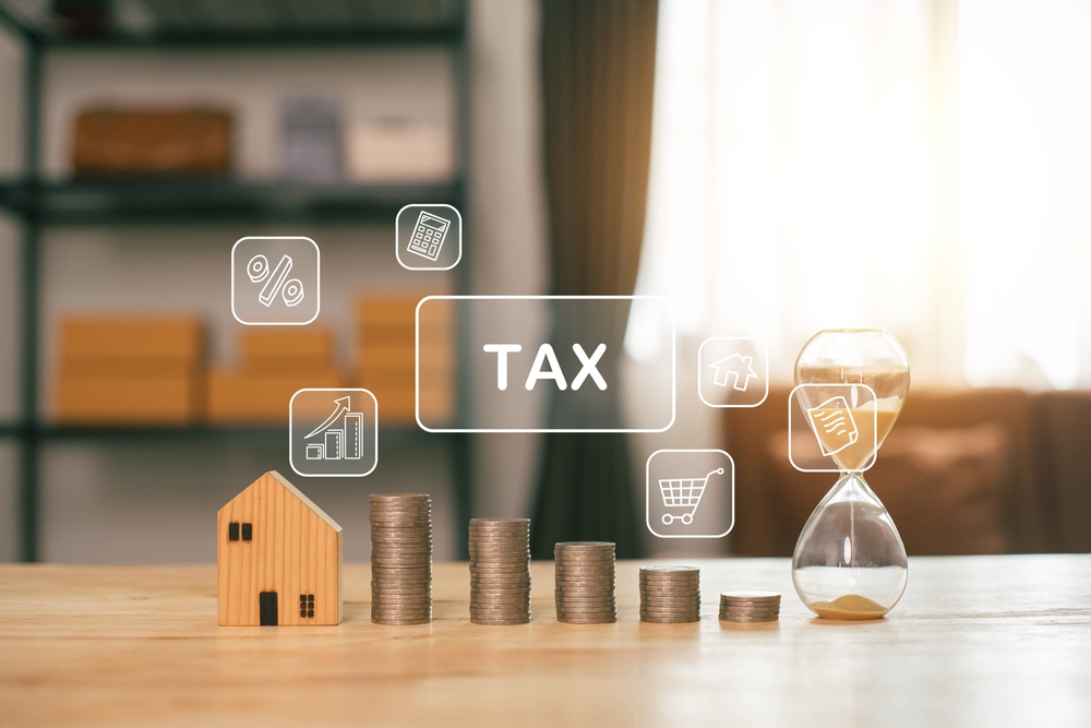 A tabletop display features a miniature wooden house, stacks of coins, and an hourglass with digital icons for taxes, homeownership, finance, and budgeting surrounding the word "TAX." The image symbolizes tax planning, wealth management, and financial decision-making.
