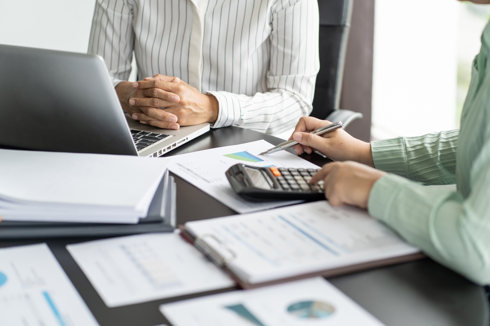 Two professionals review financial documents at a desk with a laptop, calculator, and reports during a business meeting.